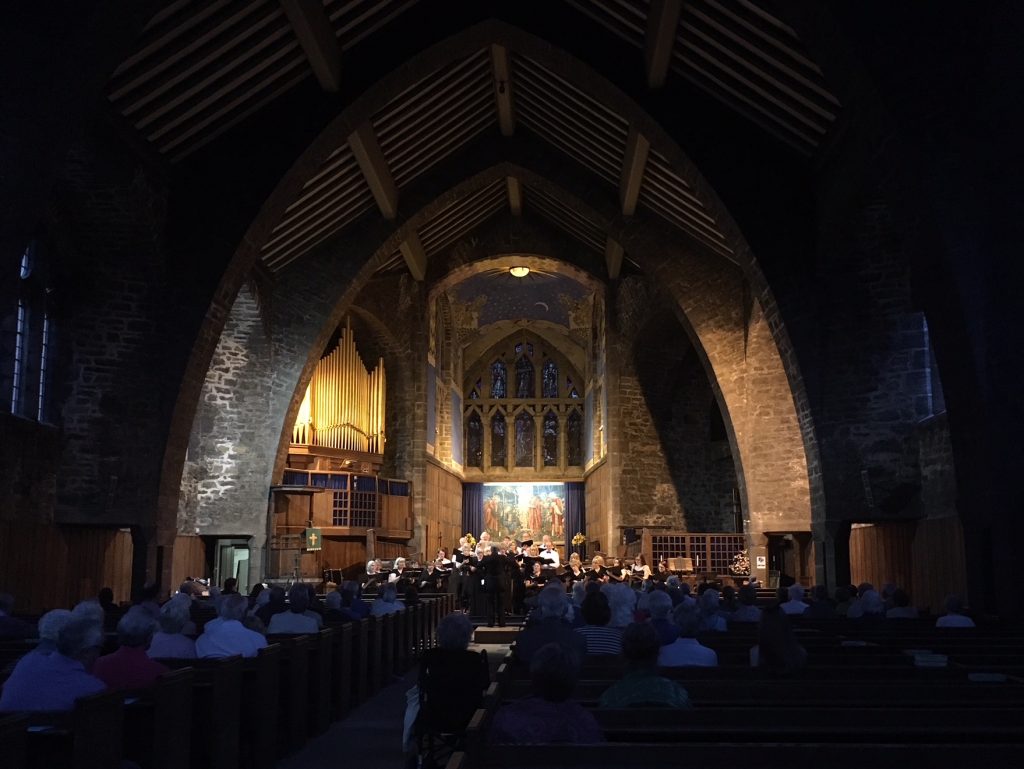 Choir in St Andrew's Church. A large choir dressed in black are stood towards the altar of the church. The altar is lit and the nave is in darkness.